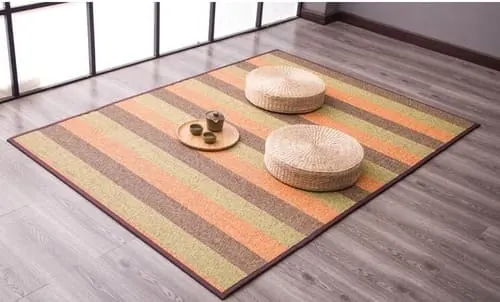 Alfombra de bambú a rayas verdes, naranjas y marrones sobre suelo de madera, con dos pufs de fibra natural