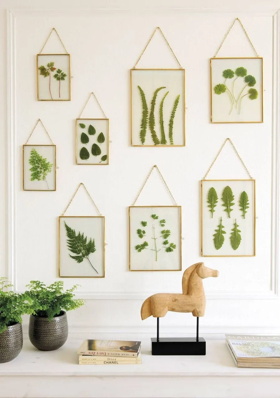 Cuadros con hojas prensadas en marcos dorados sobre una pared blanca, con plantas y un caballo de madera