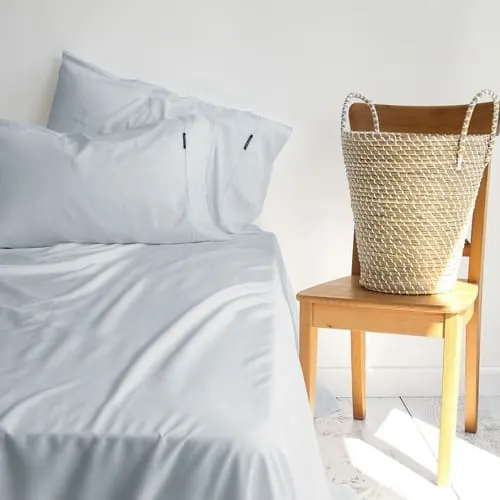 Cama con sábanas y almohadas blancas junto a una silla de madera con un cesto de fibra natural